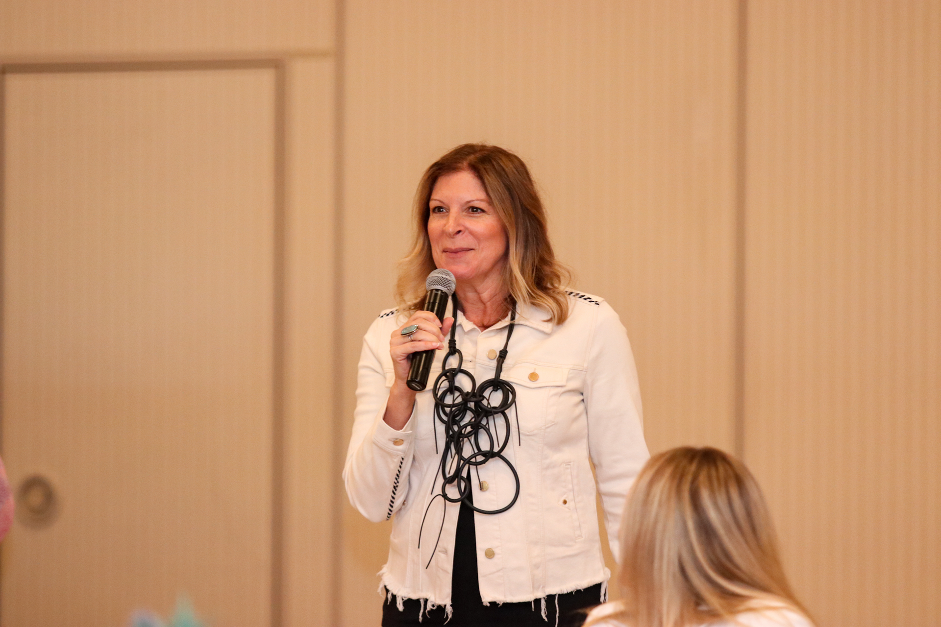 Woman in white jacket speaks into a microphone, standing in front of a neutral backdrop.