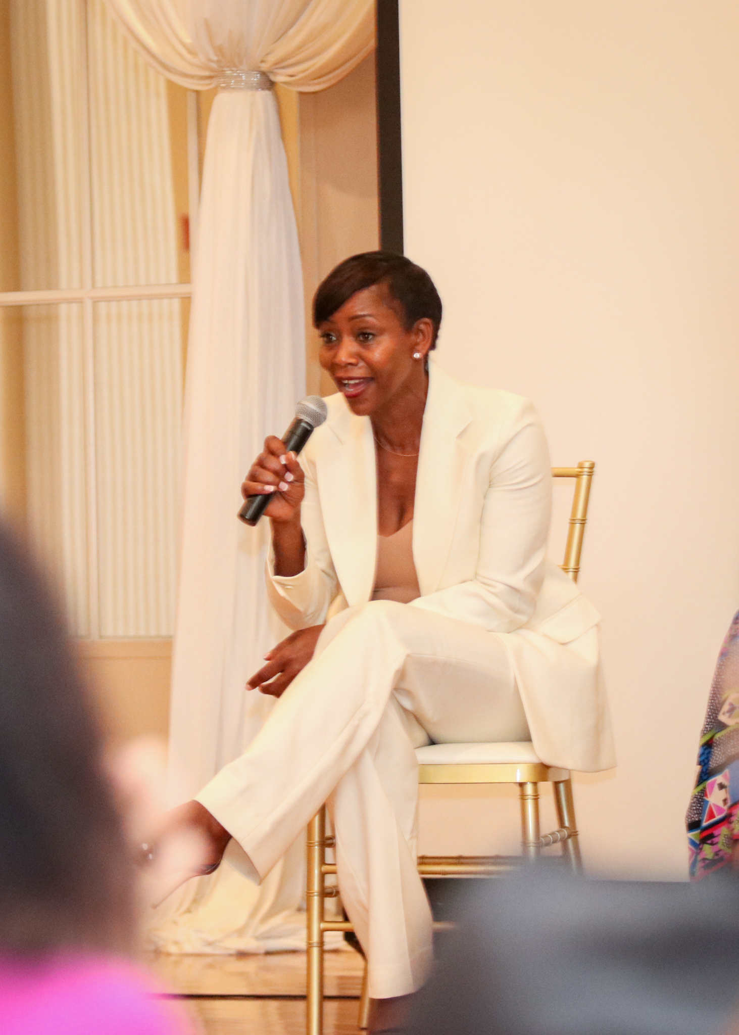 Woman in a white suit, seated and speaking into a microphone. Light-colored room, seated on a gold chair.