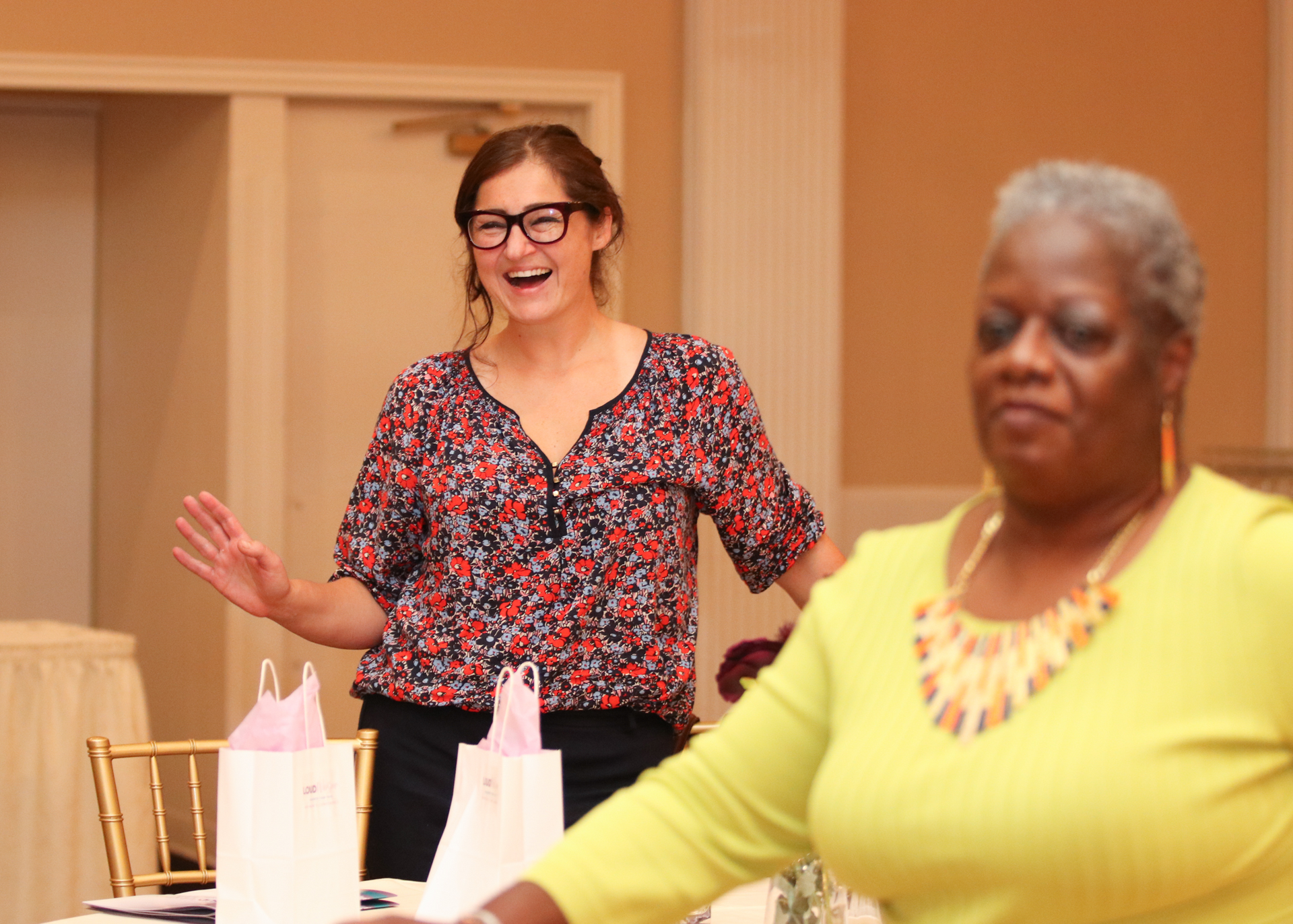 Woman in glasses laughs, arms raised, near a woman in yellow top at an event, with gift bags on a table.
