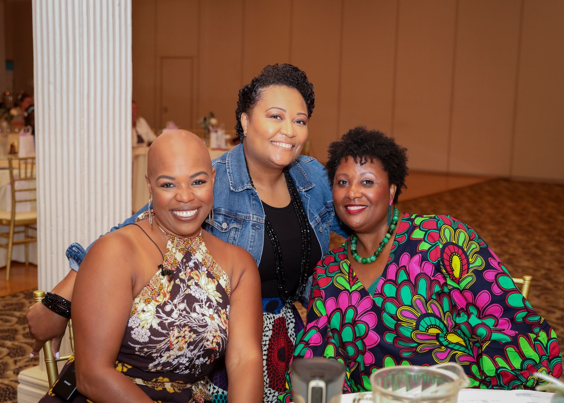 Three smiling women at a table; one bald, one in denim jacket, and one in a colorful patterned top.
