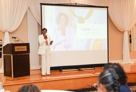 Woman in white suit gives a presentation on stage. Projector screen shows her image.