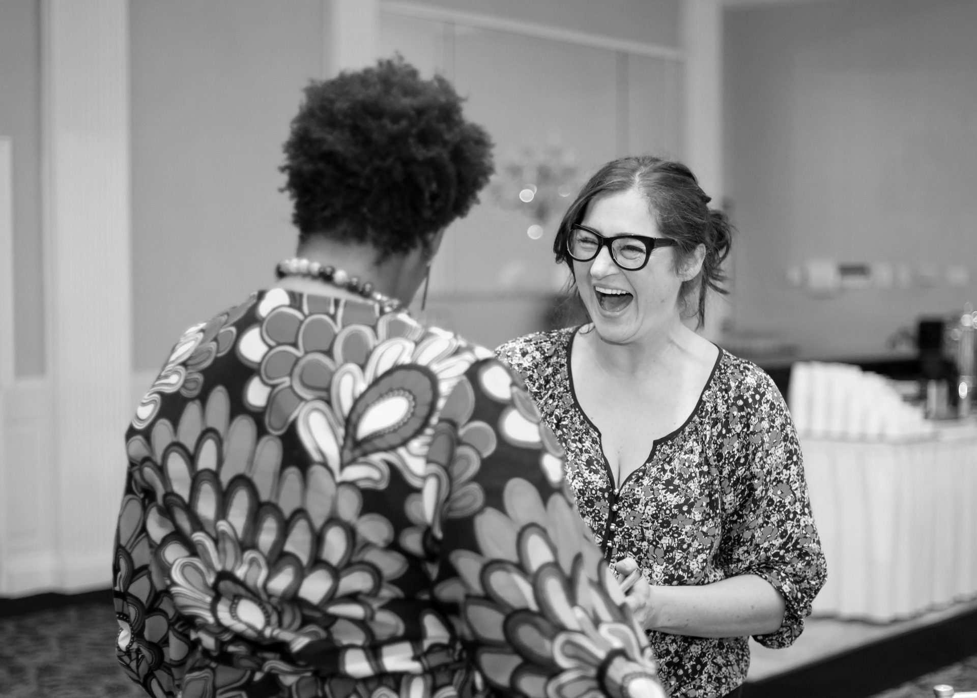 Two women laughing, one wearing glasses and a floral top, the other in a patterned shirt, inside a room.
