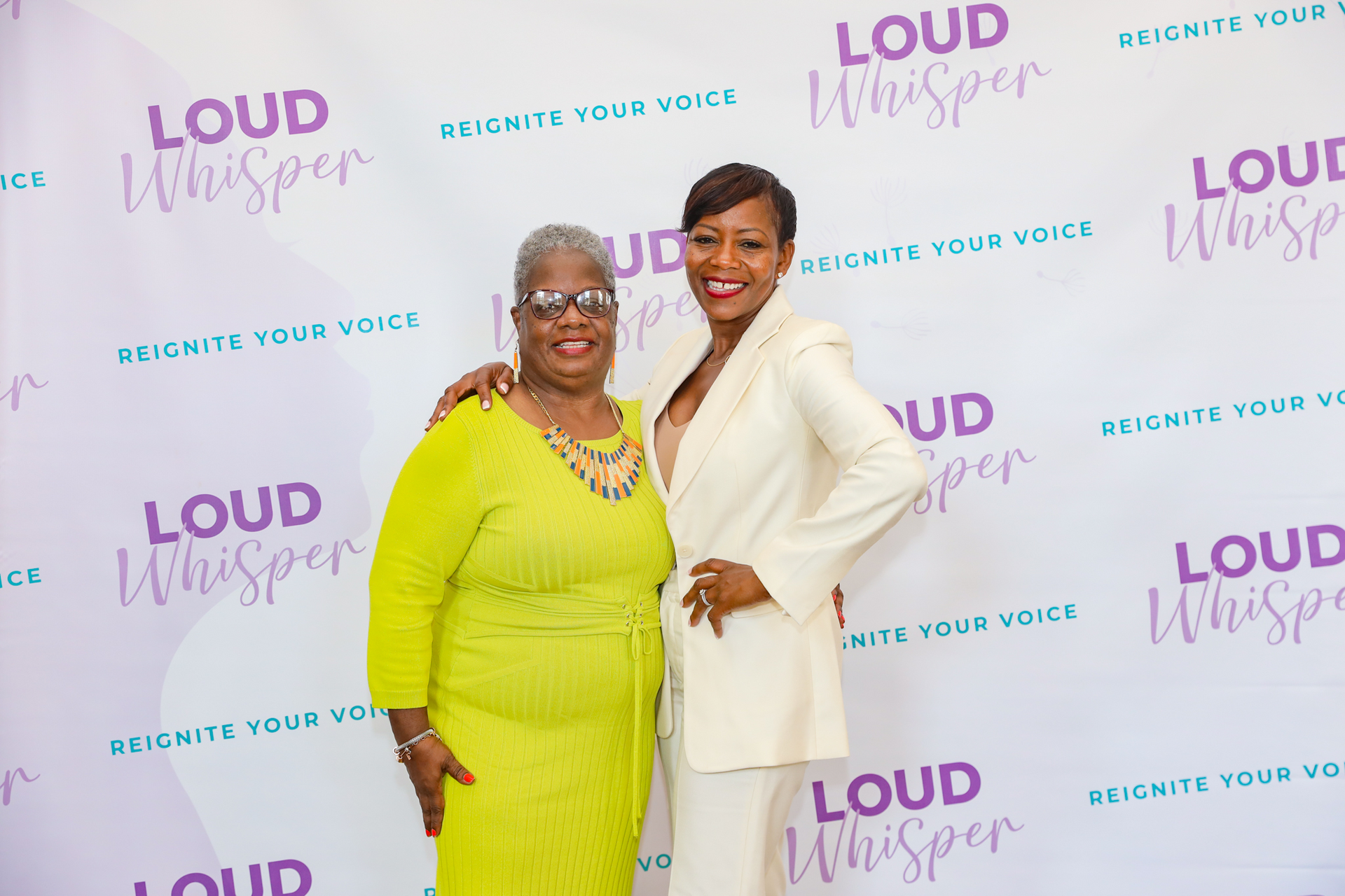 Two women pose in front of a backdrop with 