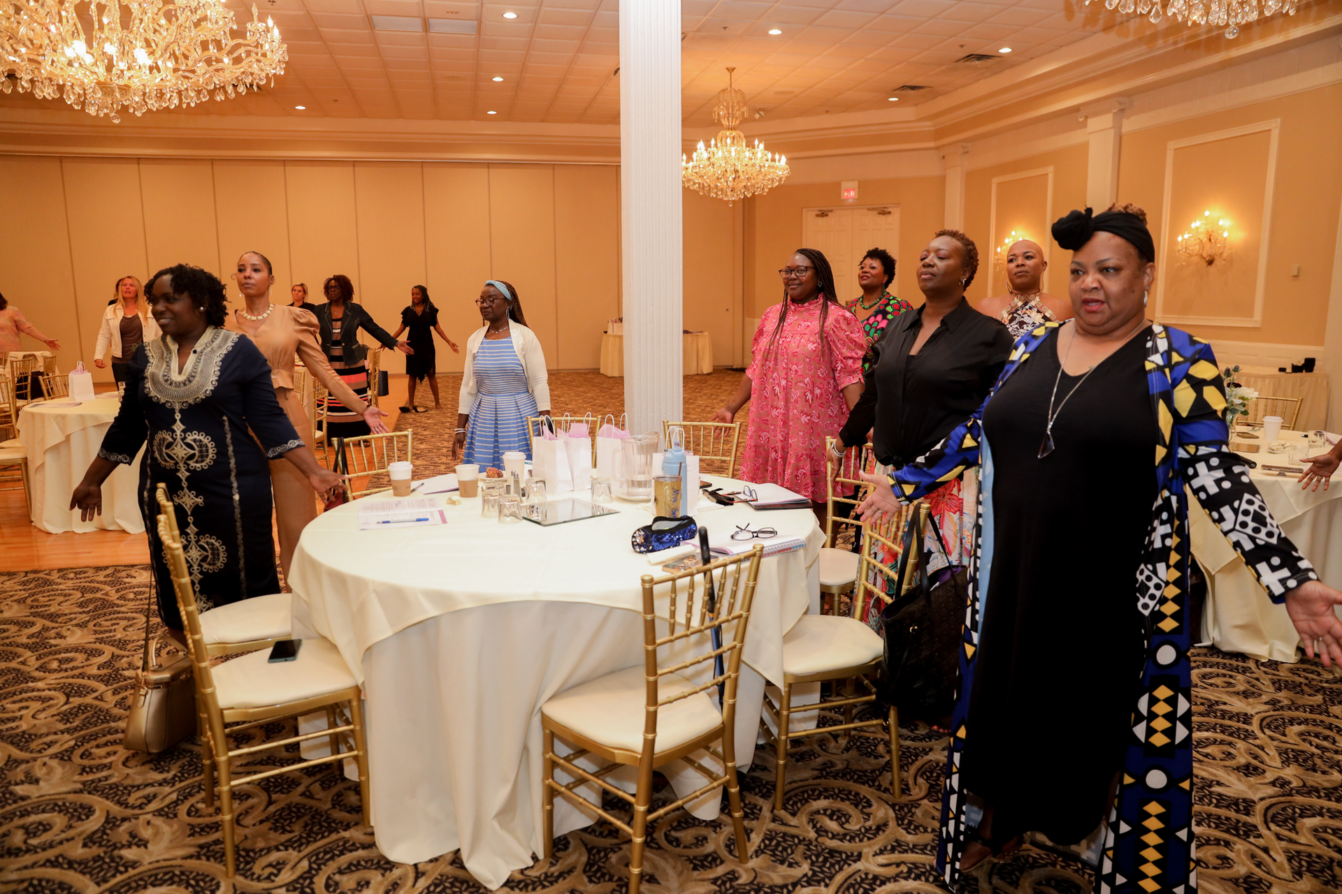 People in a ballroom with arms outstretched, around a table, and near gold chairs, possibly dancing.