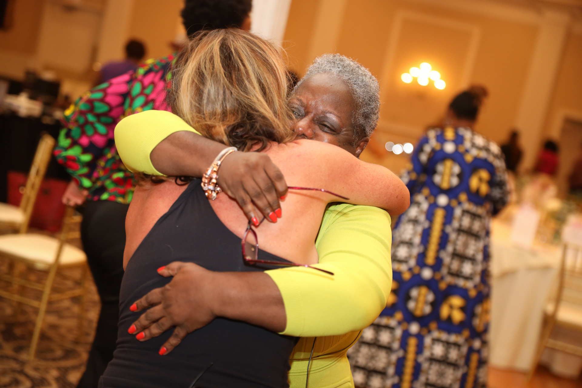 Two women embracing in a brightly lit room with a background of other people; one in yellow and the other in black.