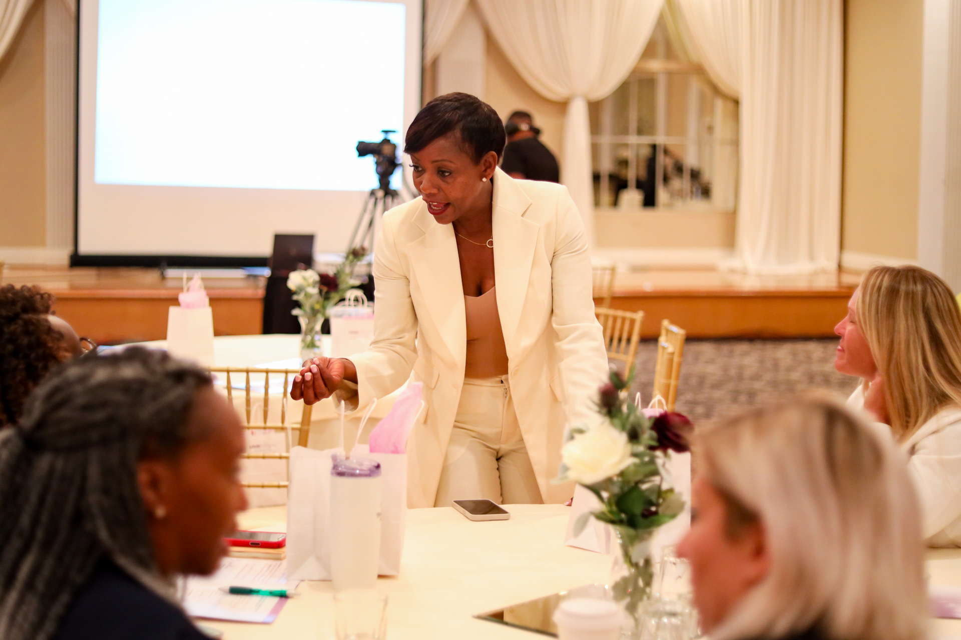 Woman in cream suit speaks at a table to other women in a brightly lit room with a projector screen.