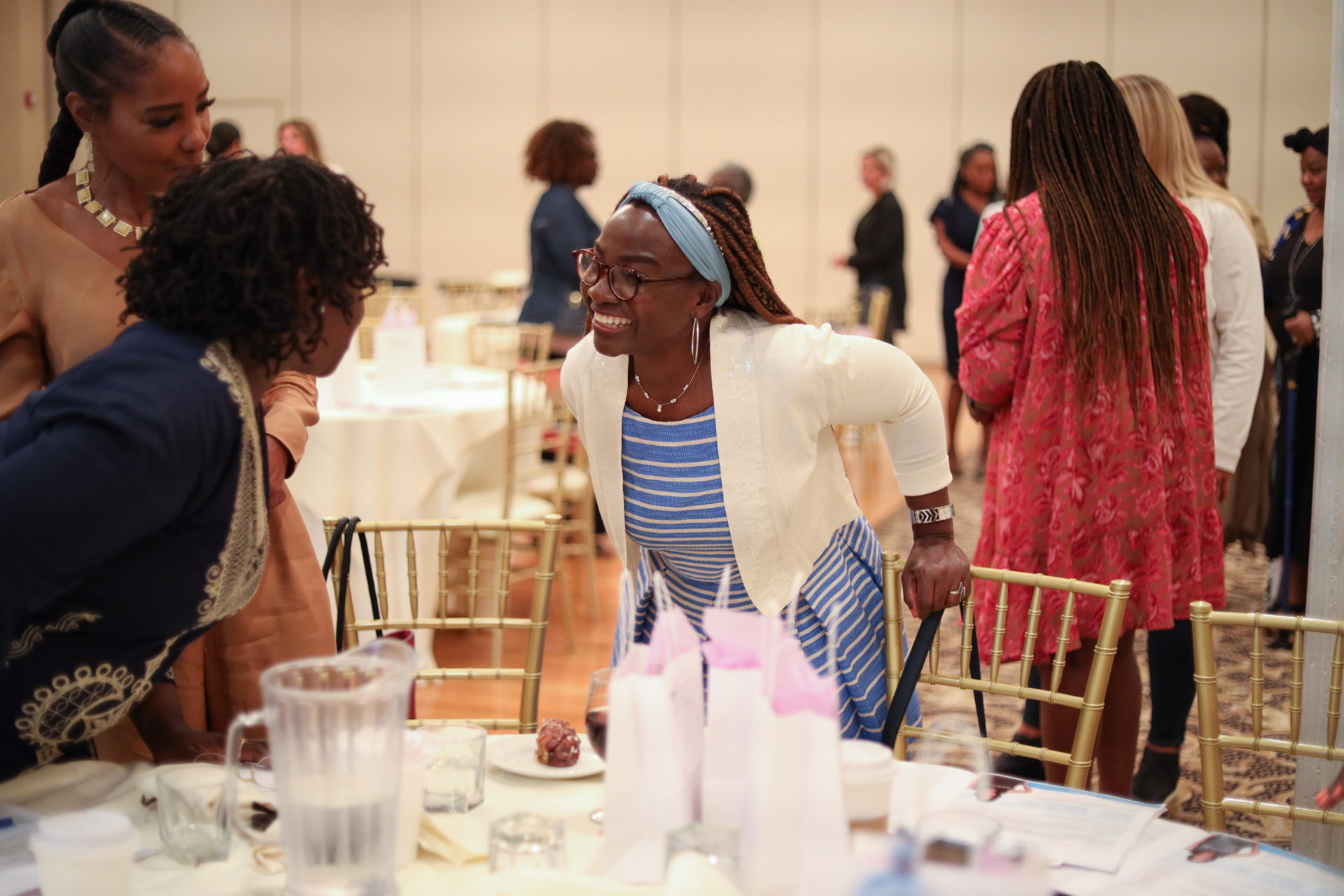 Two women in animated conversation at a table; others in background at event.