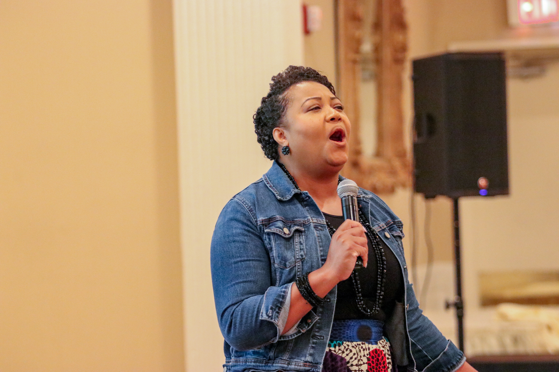 Woman singing into a microphone, wearing a denim jacket, indoors near a speaker.