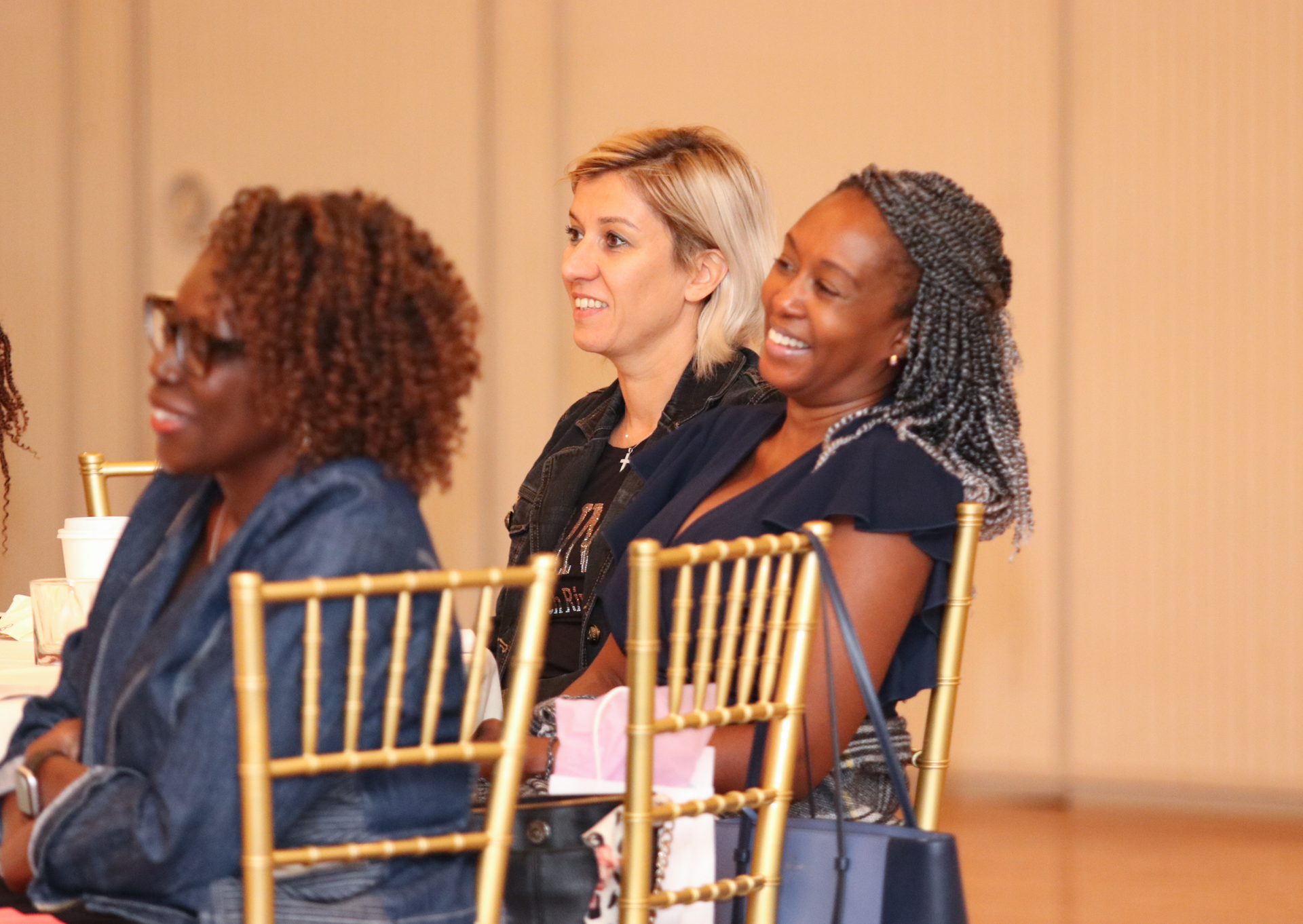 Three women smiling, seated at a table with gold chairs.