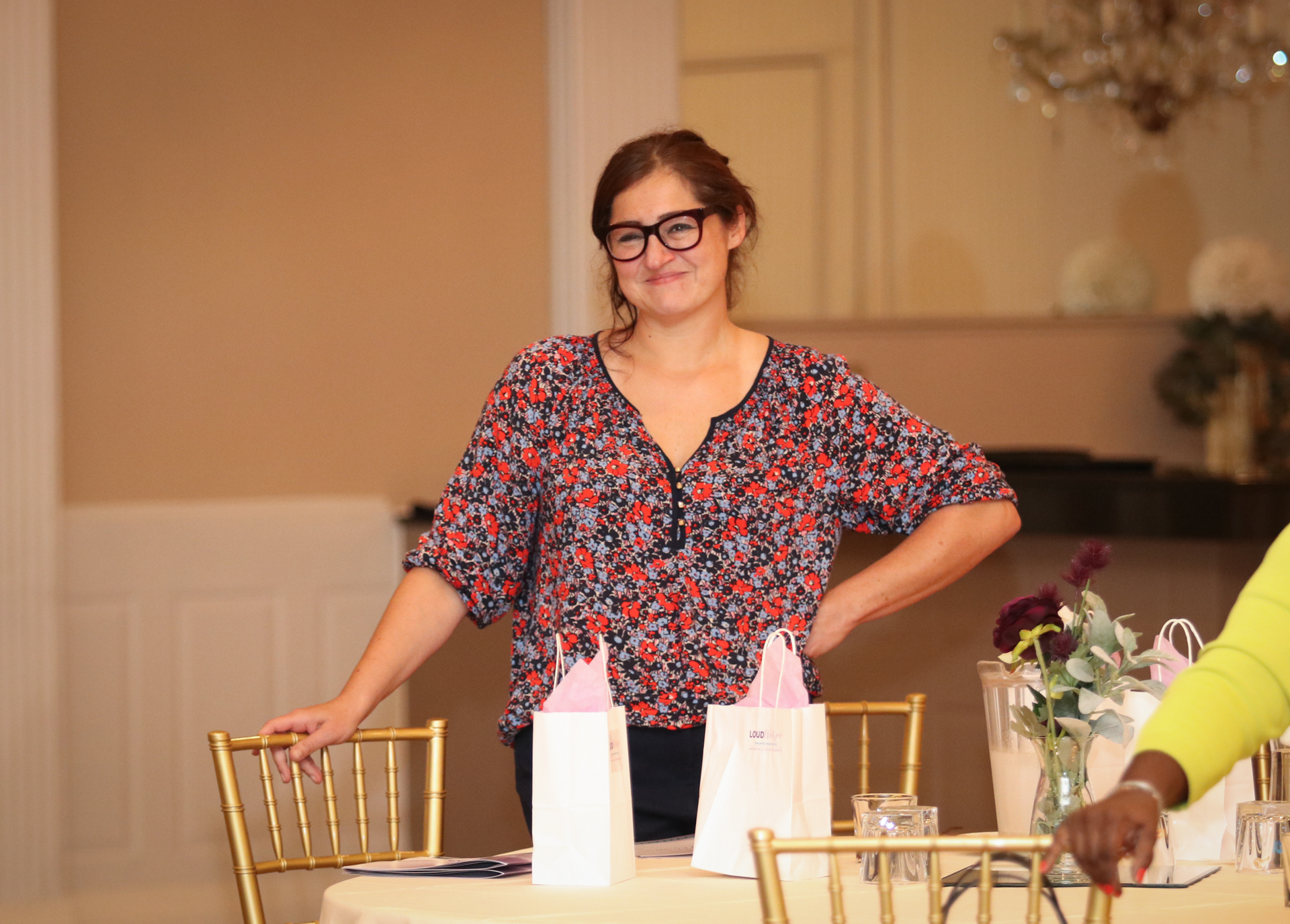 Woman with glasses smiles, standing behind a table, with gift bags, in a decorated room.
