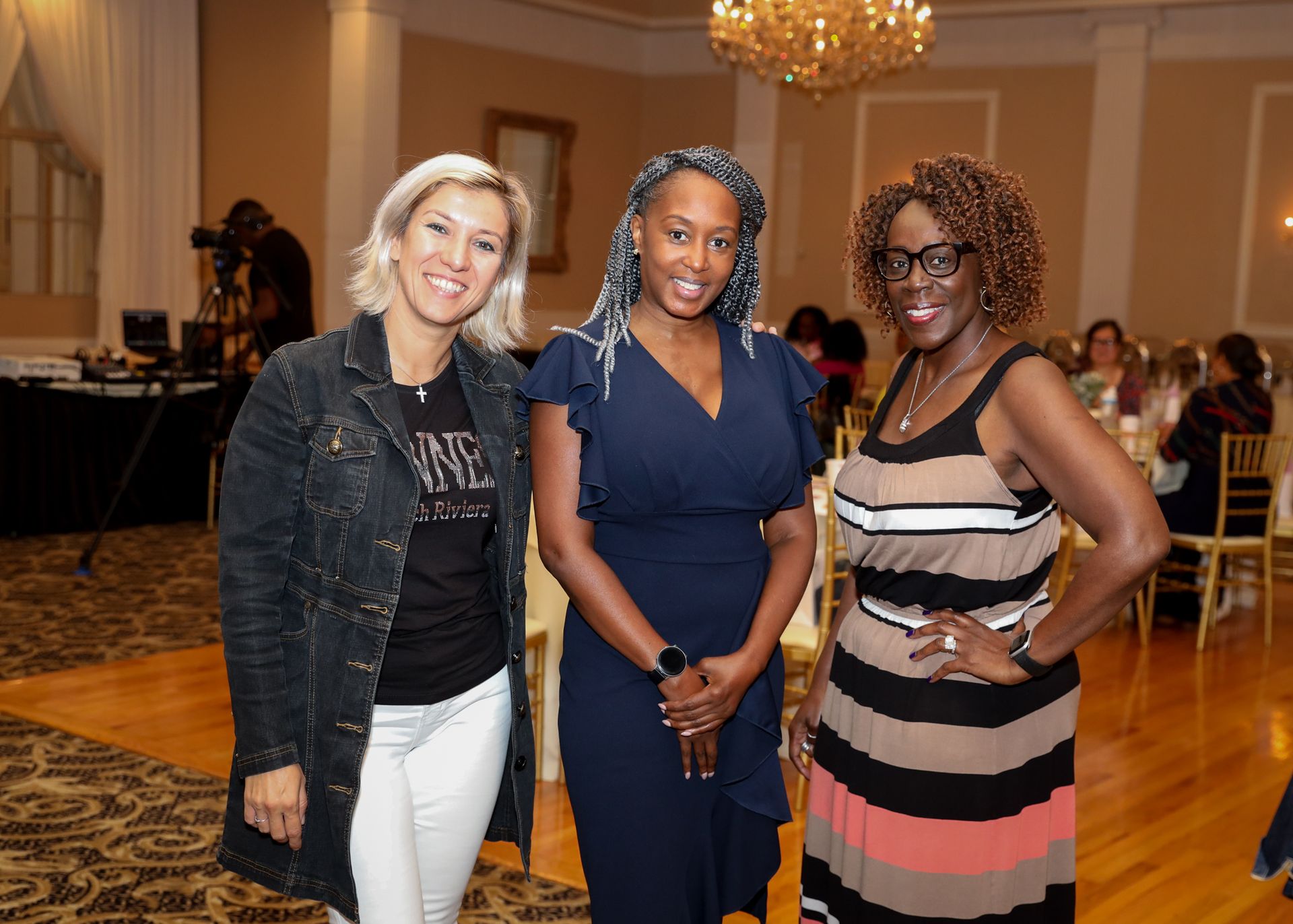 Three women smiling together in a ballroom; one in a denim jacket, one in a navy dress, and one in a striped dress.
