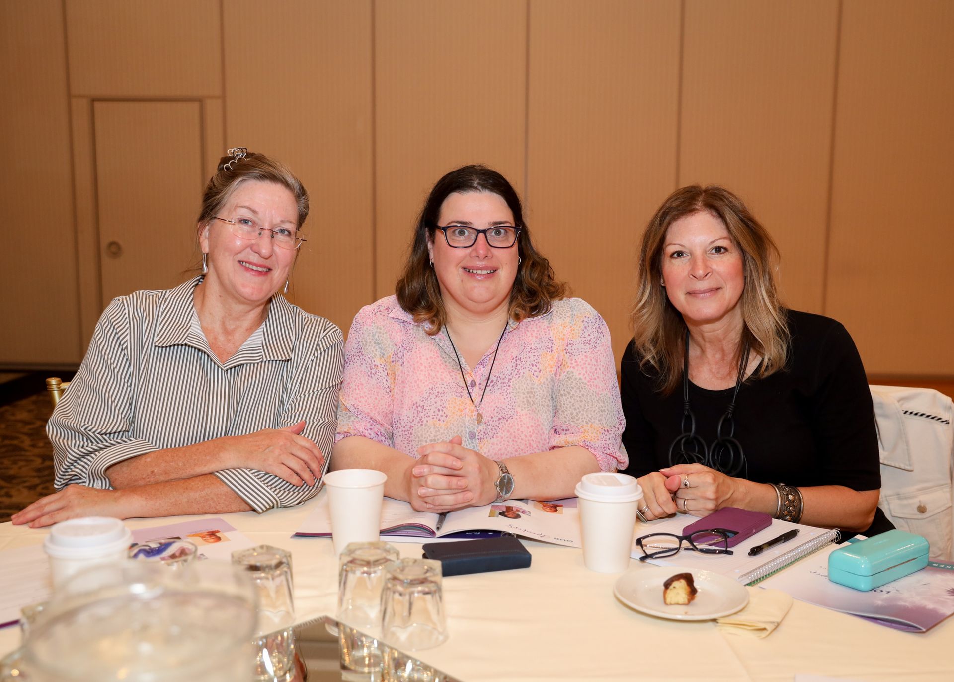 Three women seated at a table, smiling, with papers, cups, and glasses present.