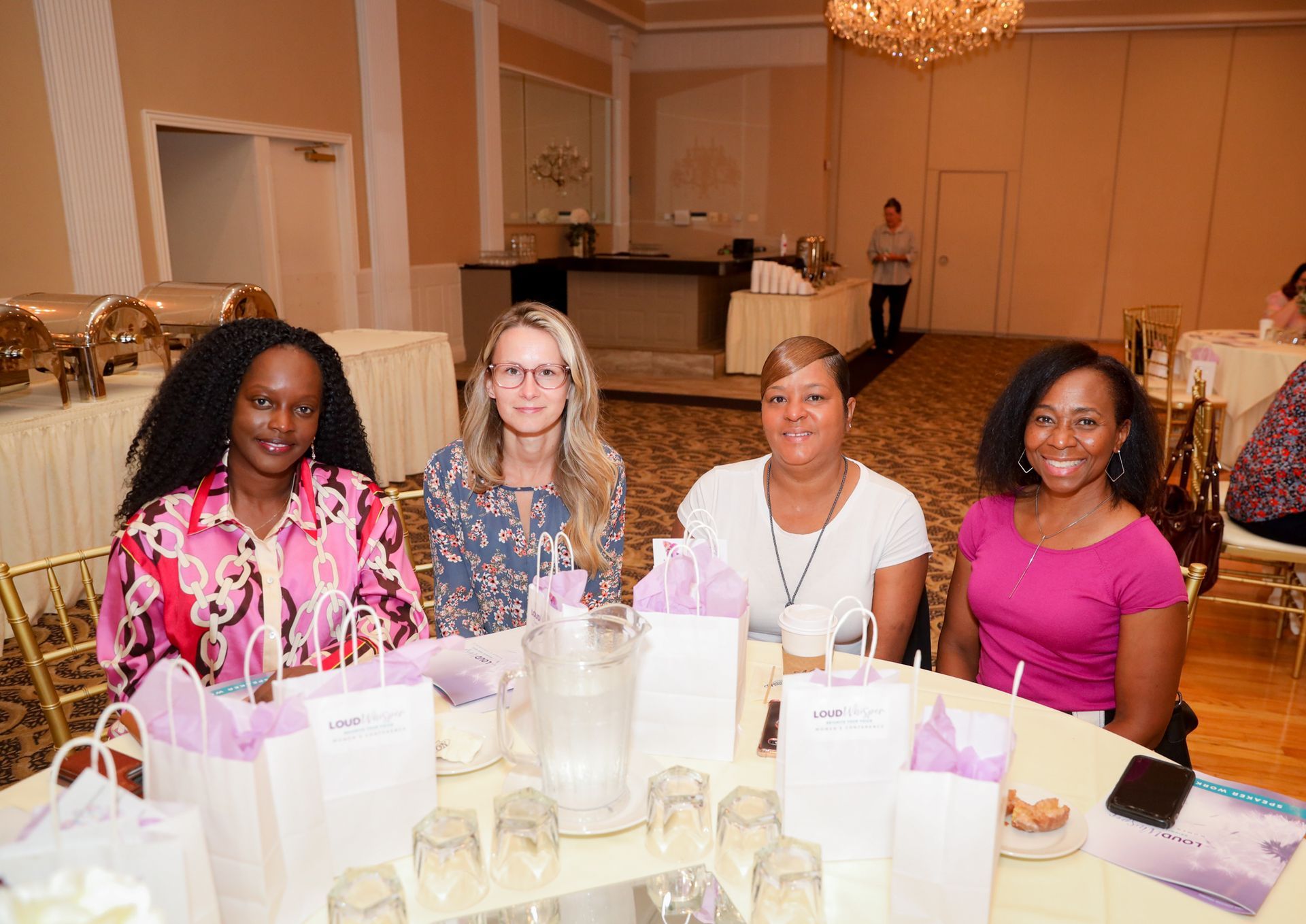Four women seated at a table in a banquet hall. They smile, with gift bags in front of them.