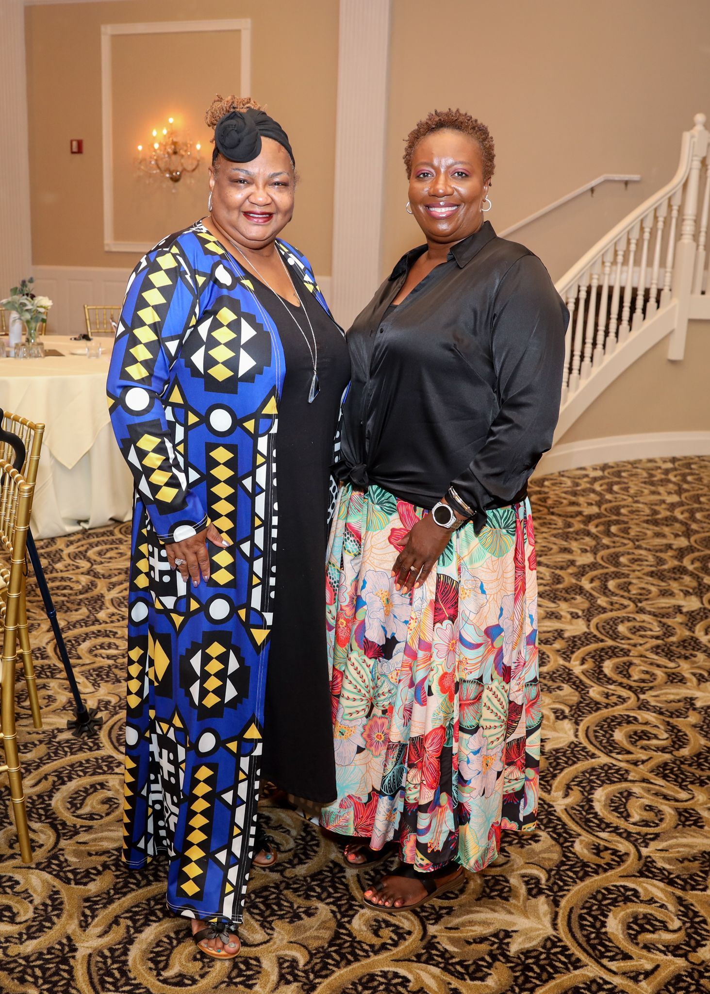Two women smiling, posing for a photo. One wears a long blue patterned coat, the other, a black top and floral skirt.