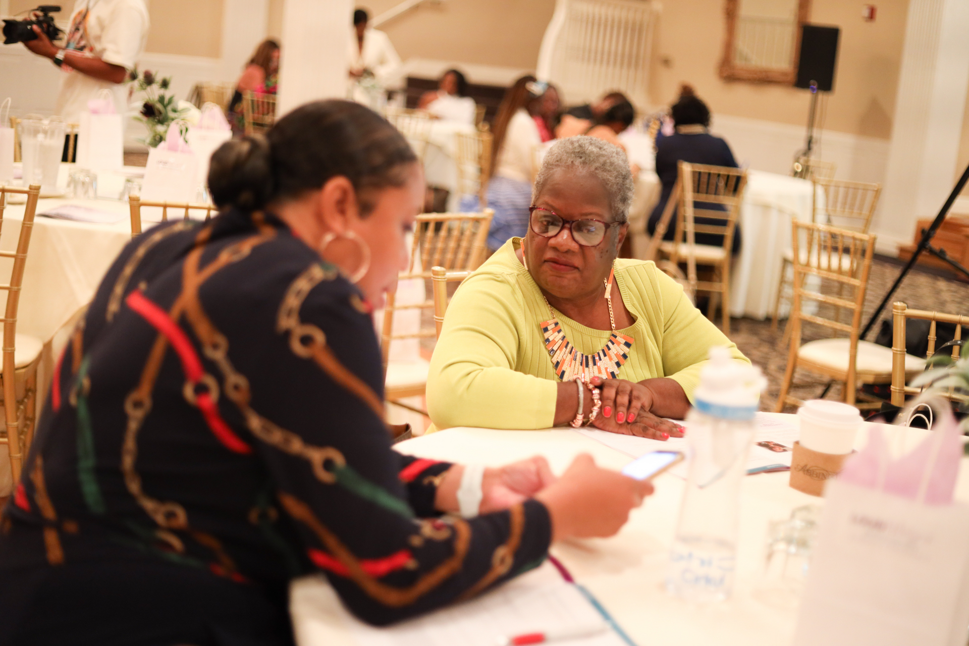 Two women at a table in a well-lit room, conversing. One wears a patterned shirt, the other a yellow sweater.