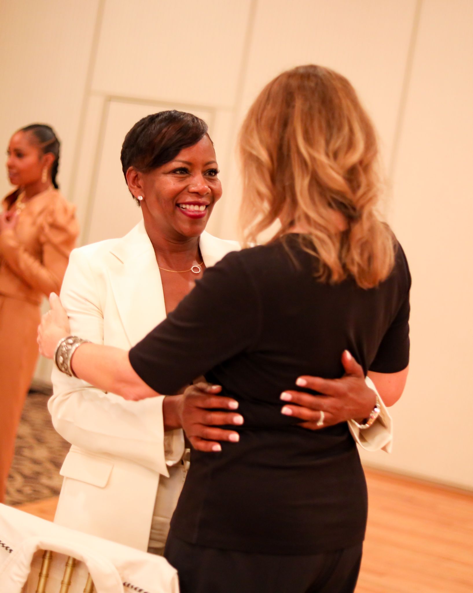 Two women embrace, smiling. One in white jacket, the other in black top. Another woman in background applauds.