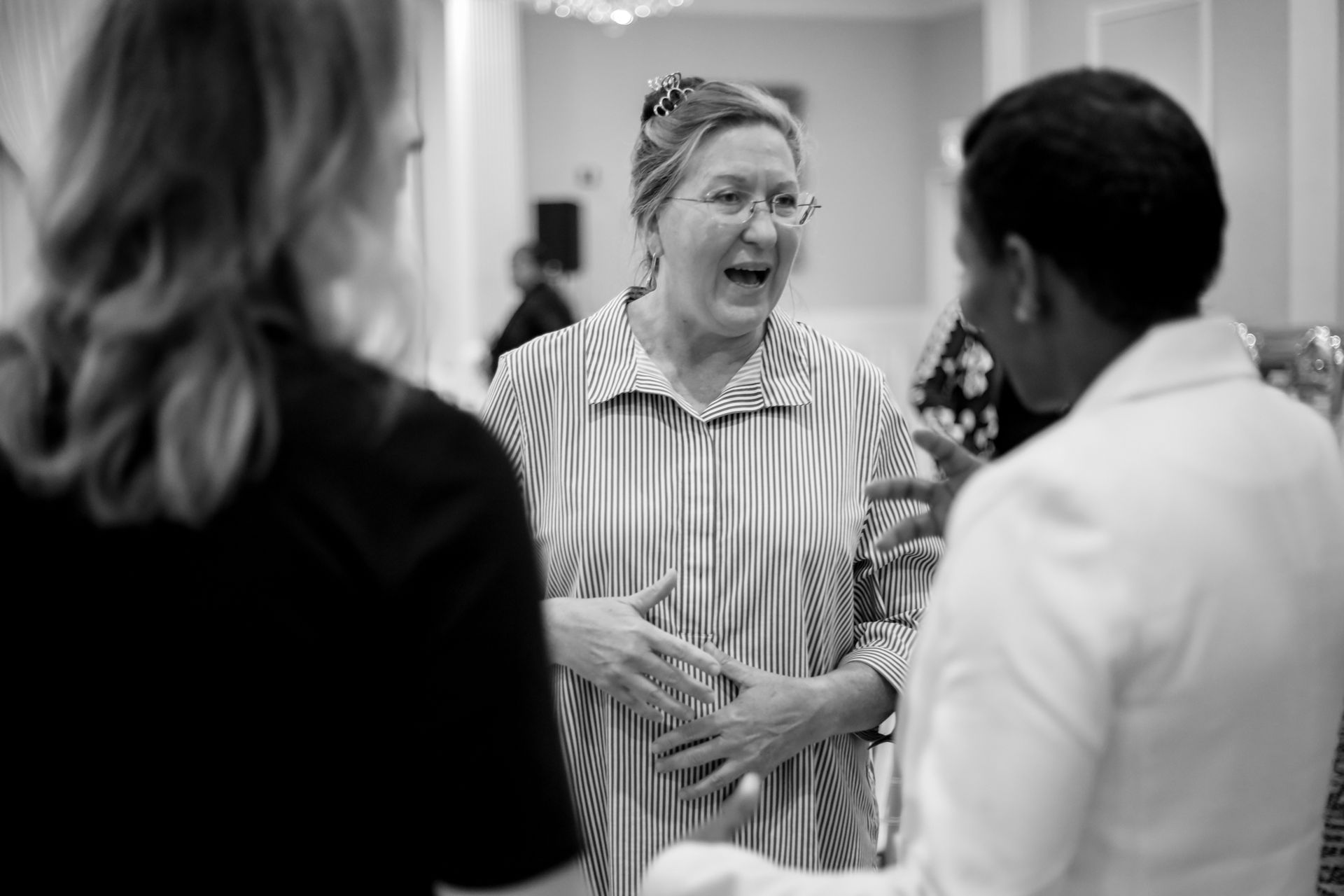 Three people in conversation, indoors. Woman in striped shirt speaks with hand gestures; two others face her.