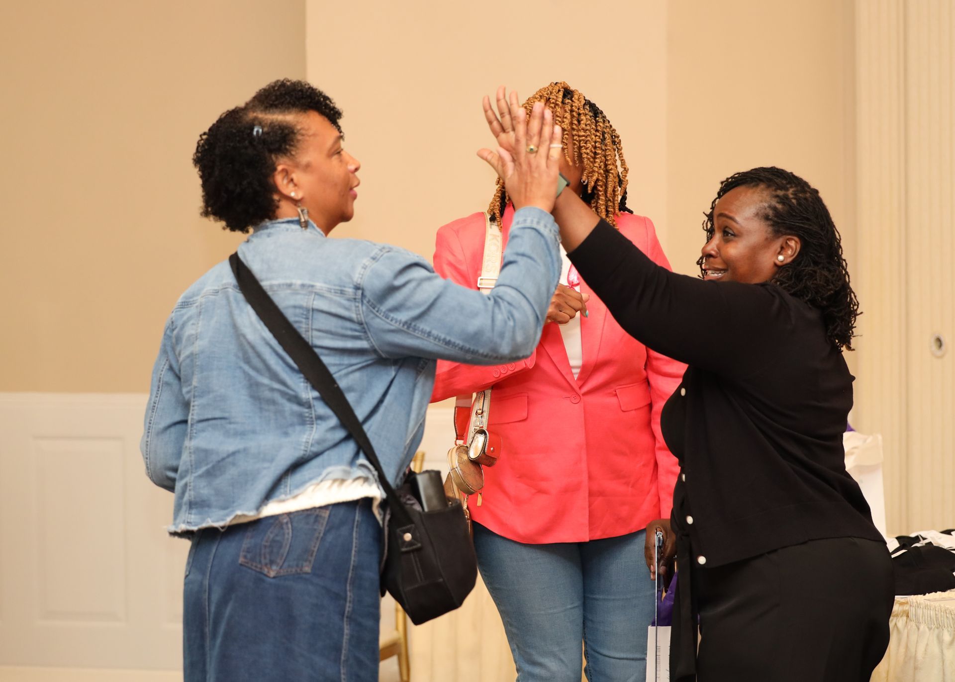 Three people indoors: two women high-fiving, the third woman in between. One wears a denim jacket, another a black blazer.