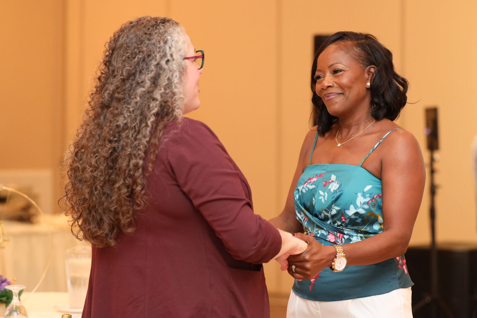 Two women holding hands, smiling, in a ballroom. One has long gray hair, the other wears a teal top.