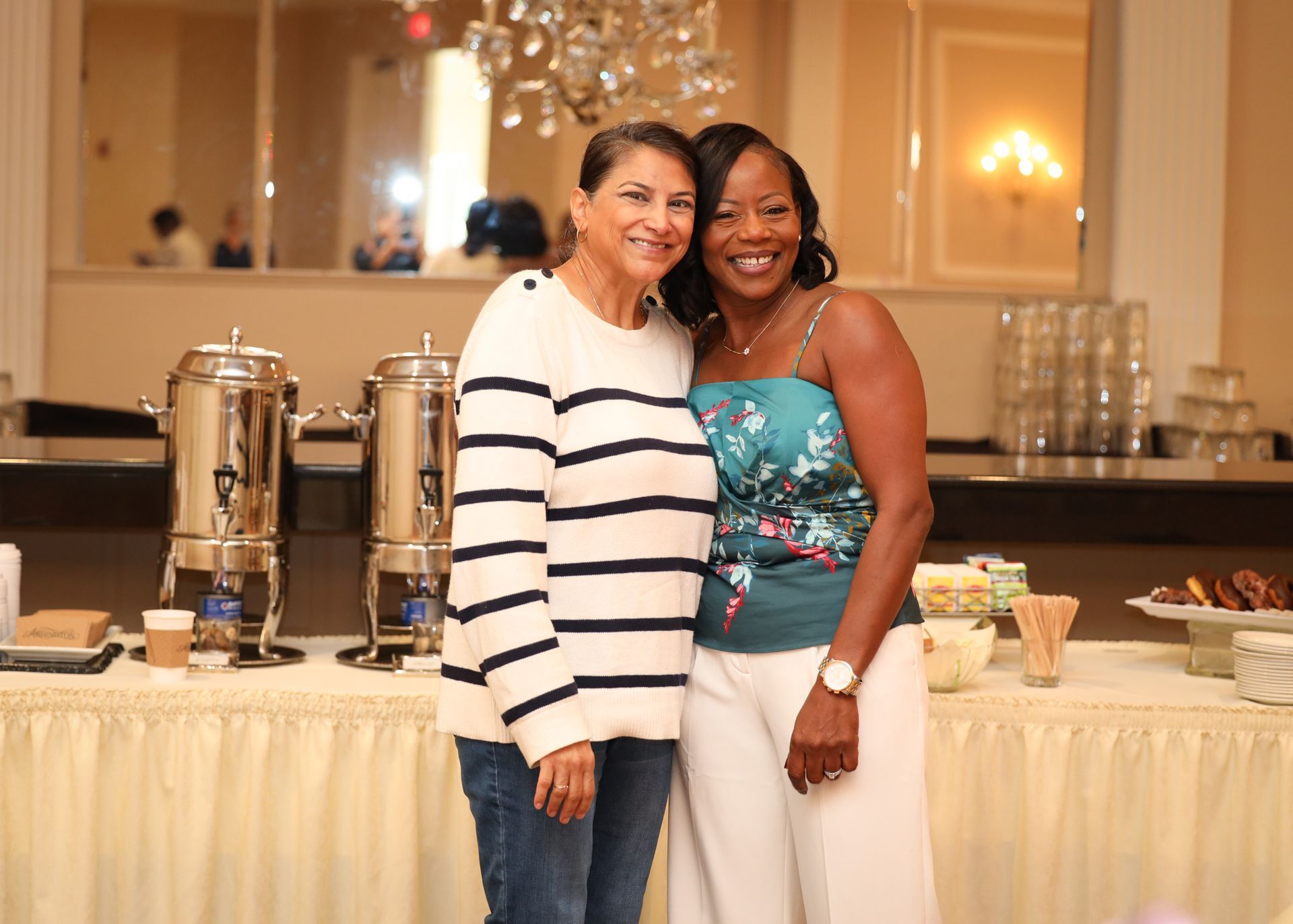 Two women smiling, posing together behind a buffet table with coffee urns, pastries, and a chandelier.