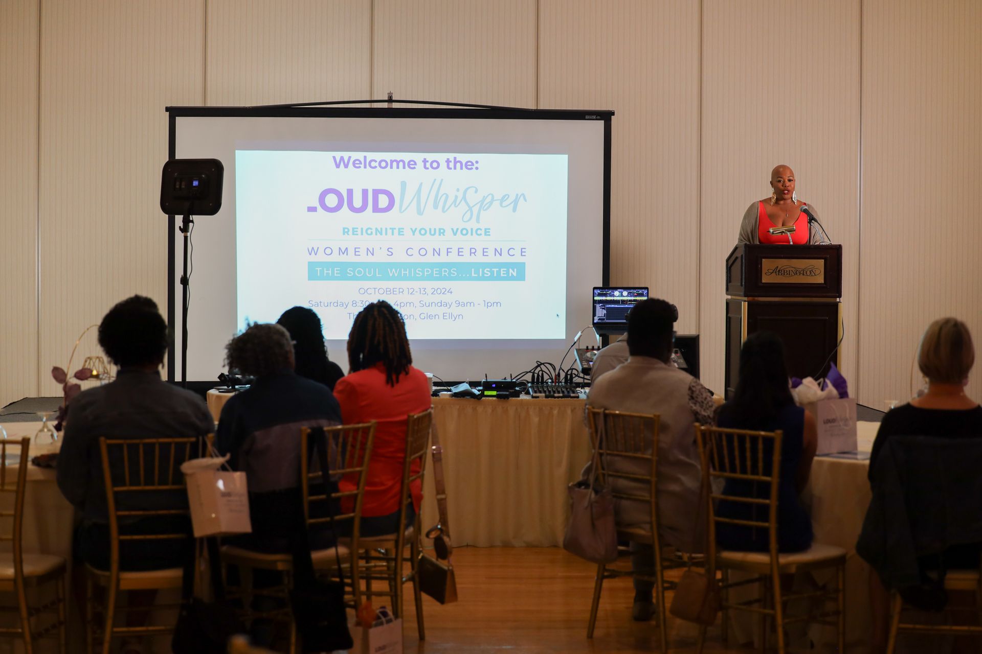 A speaker on a podium at a conference, presenting to a seated audience, with a projected screen behind them.