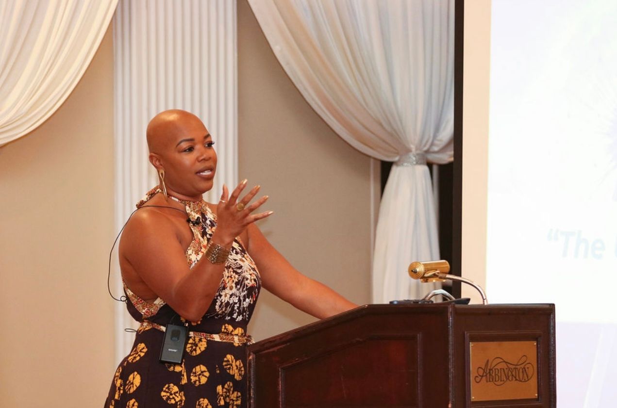 Woman with shaved head giving a presentation at a podium. She gestures with hands. Beige and white backdrop.