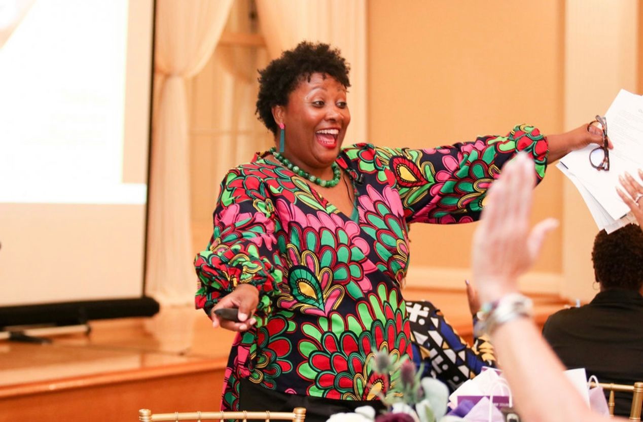 Woman in colorful top, gesturing enthusiastically while speaking at an event; hands raised in response.