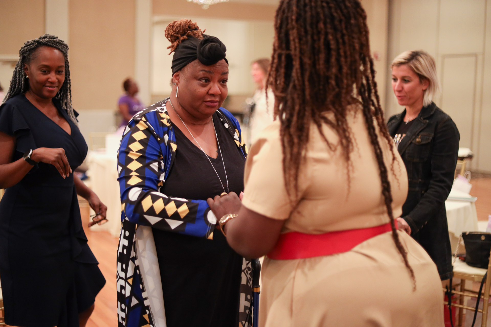 Women in a well-lit room converse, some gesturing. One wears a blue patterned cardigan, another a red belt.