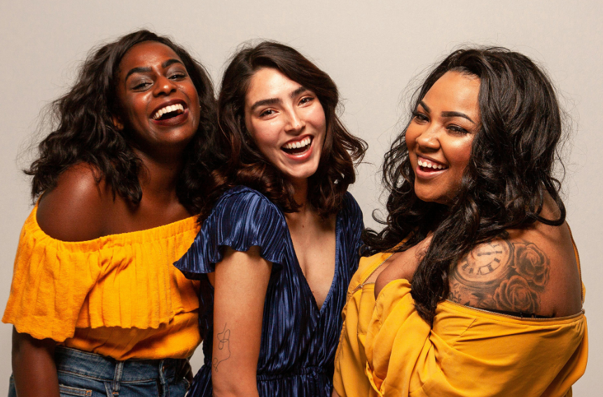 Three women laughing, embracing. Two wear yellow off-shoulder tops, one wears a blue dress. White backdrop.