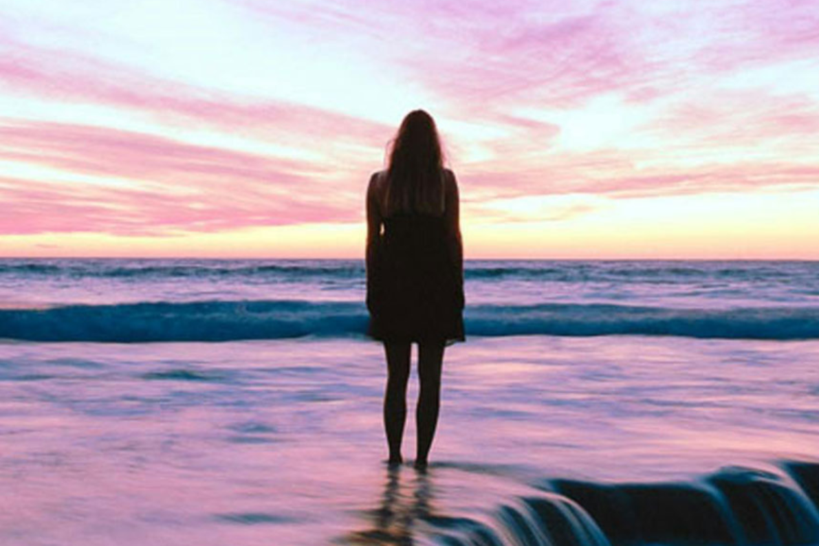 Woman standing in ocean, facing sunset with pink and yellow sky.