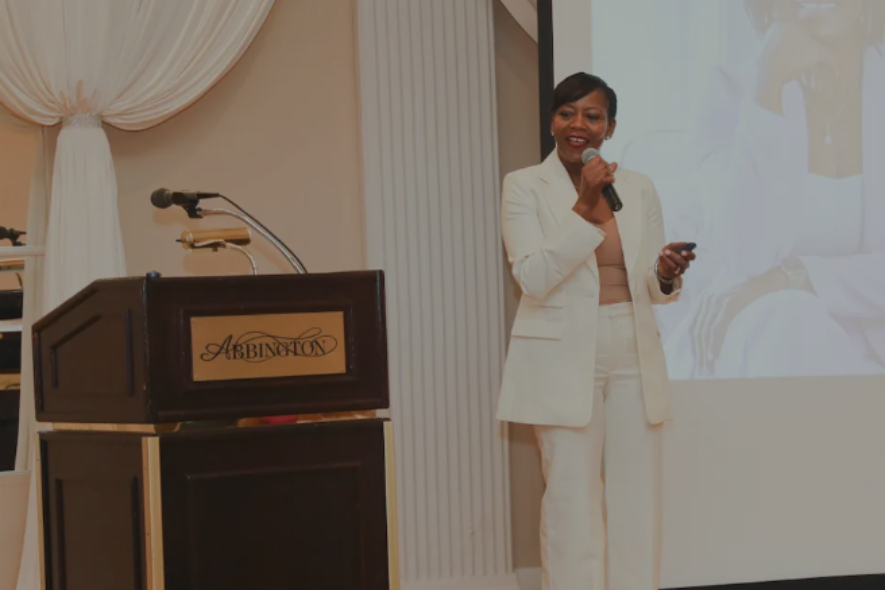 Woman in white suit speaking at a podium, holding a microphone, inside a building.