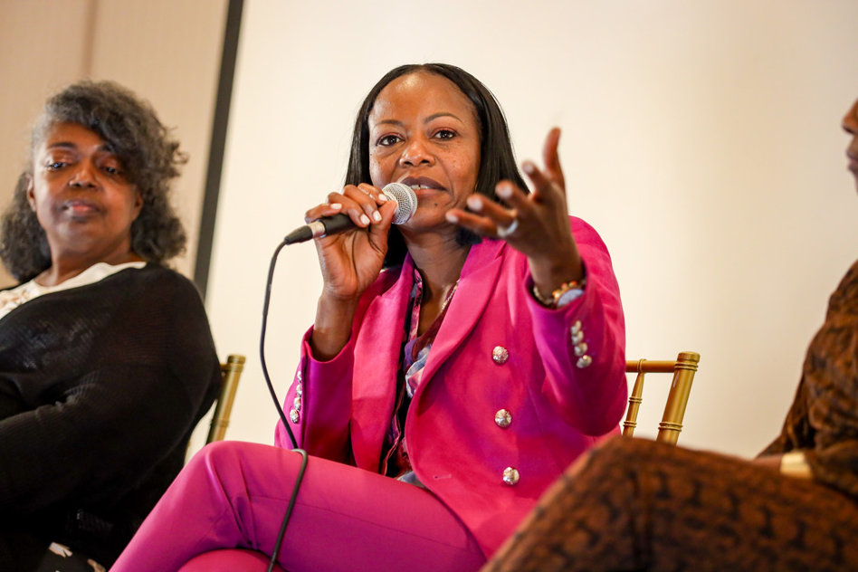 Woman in pink suit speaks into a microphone at a panel discussion. Two other women are seated nearby.