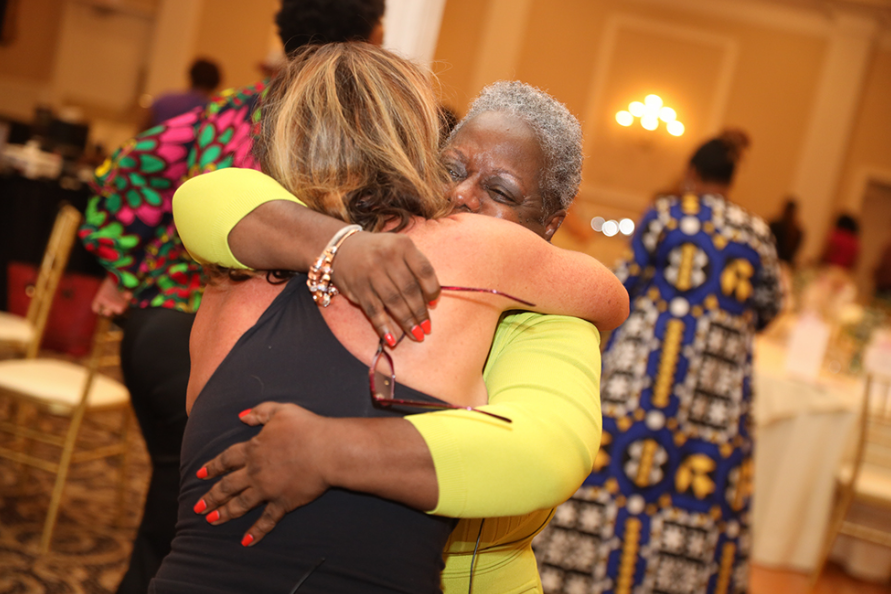 Two women embrace indoors, one in black tank top, the other in yellow. They are smiling and appear happy.