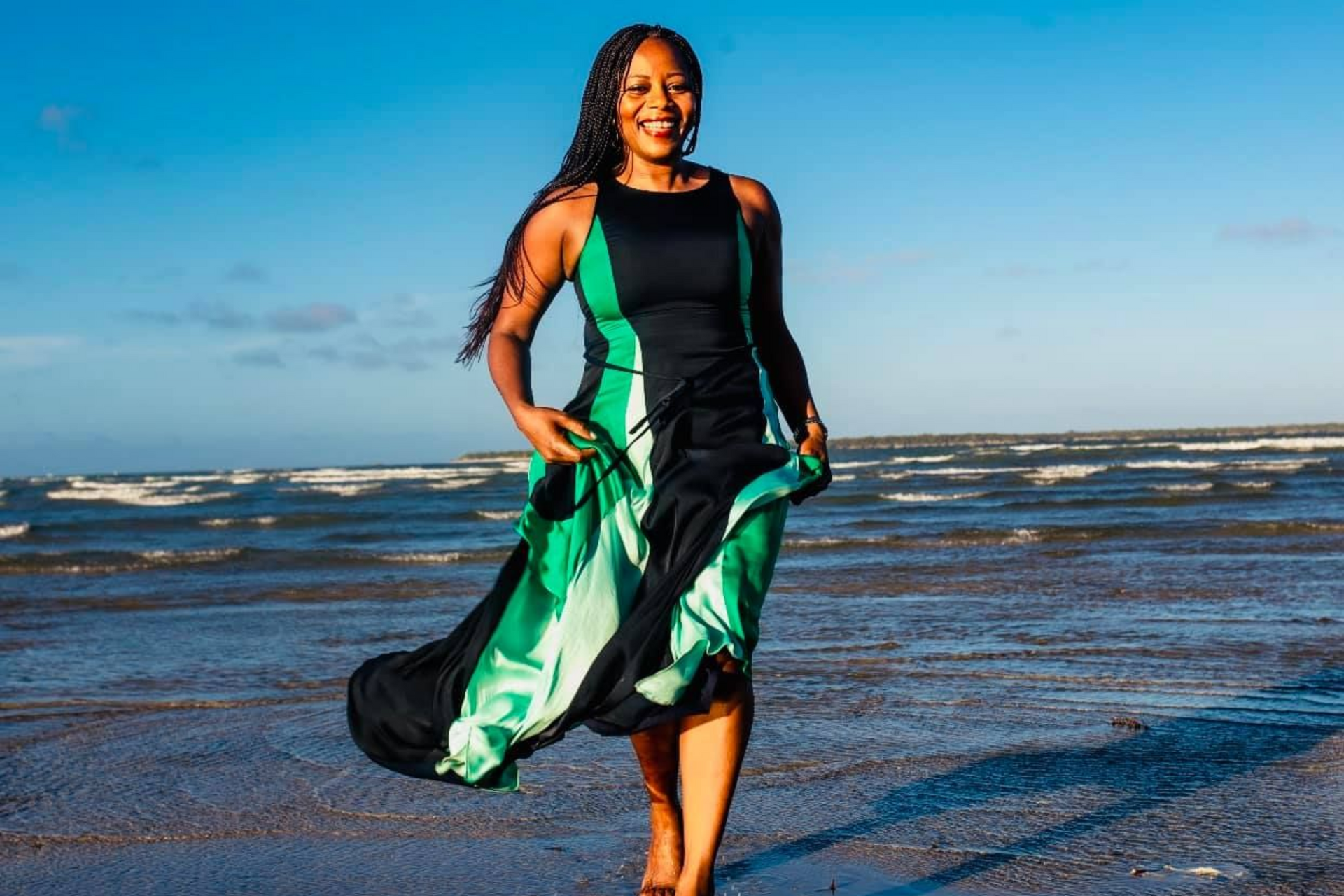 Woman in flowing green and black dress, smiling and walking on a beach near the ocean.