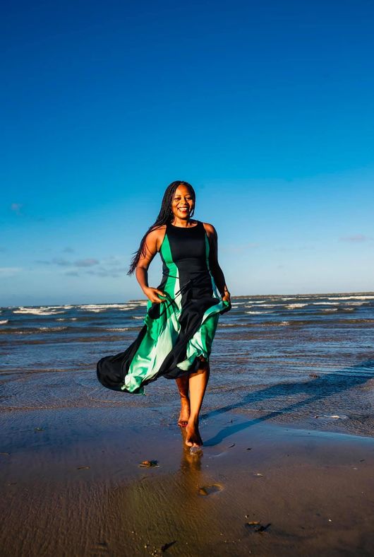 Woman in green and black dress walks barefoot on beach, waves in background.