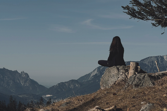 A person sits in a meditative pose on a rock overlooking a vast mountain landscape under a clear sky.