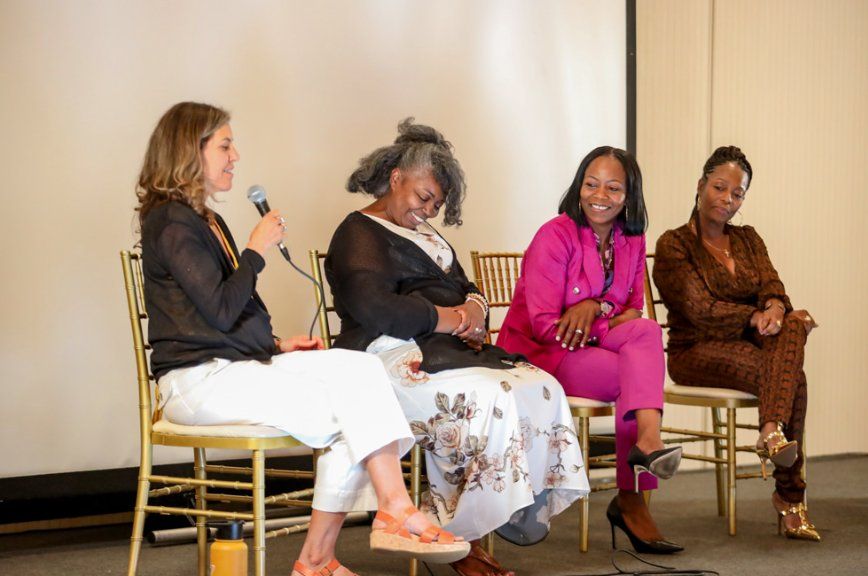 Four women sit on chairs on a stage. One holds a microphone, speaking. The others listen and smile.