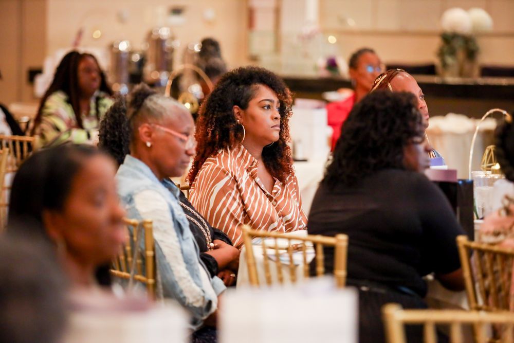 People seated at tables in a brightly lit room, some looking forward, likely at an event.