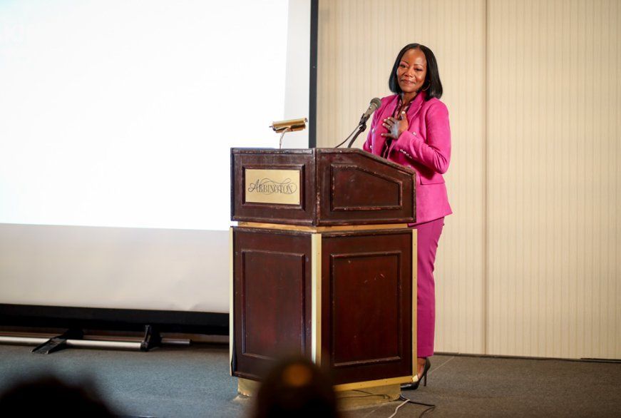 Woman in pink suit stands at a podium, speaking. Presentation screen behind her. Beige wall background.