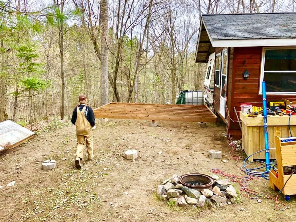 A man is standing in front of a wooden house next to a fire pit.