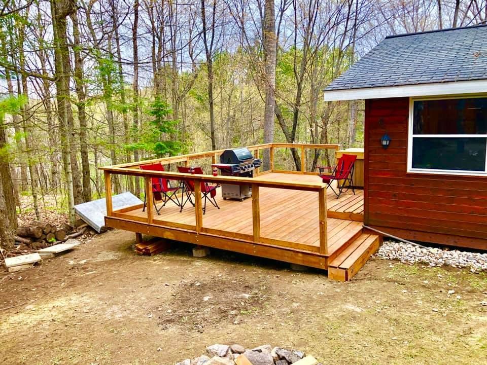 A small cabin with a wooden deck and a grill in the woods.