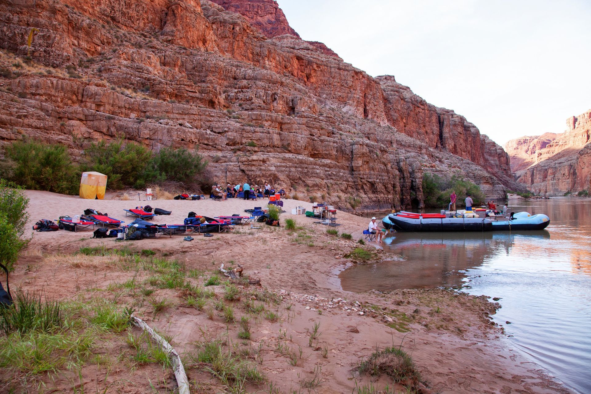 Rafts and gear on a sandy shore of a river, beside a high, reddish canyon wall.