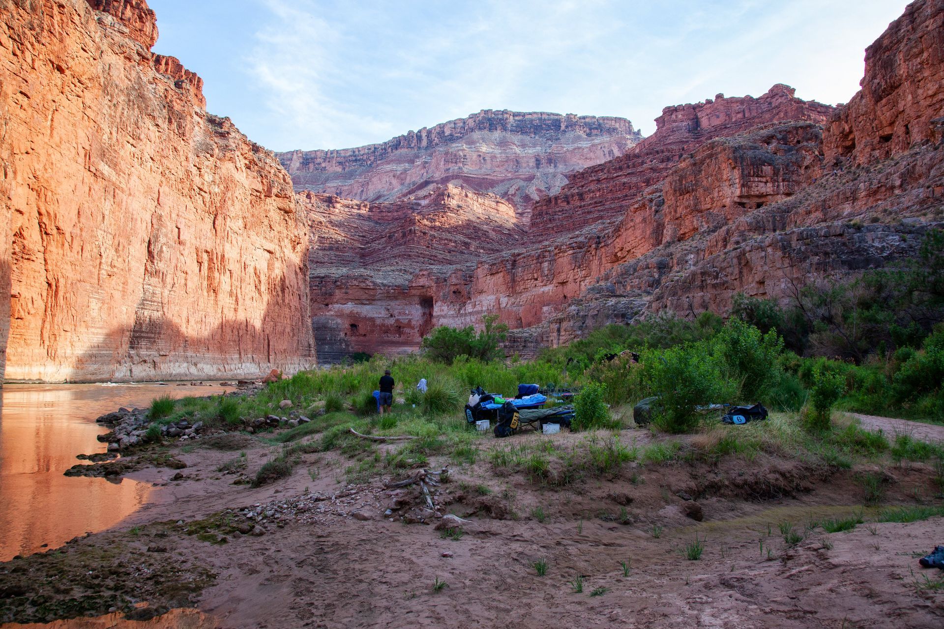 Red rock canyon with river and campsite. People gather near gear by the green bushes; the sky is blue.