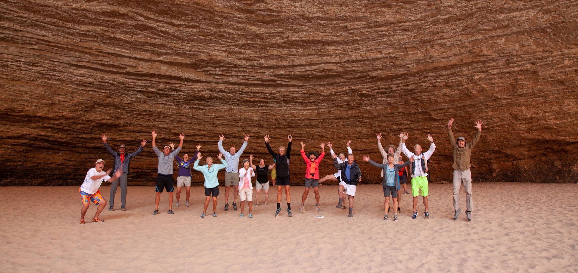 Group of people with arms raised in a cave, sandy floor, and brown rock ceiling.