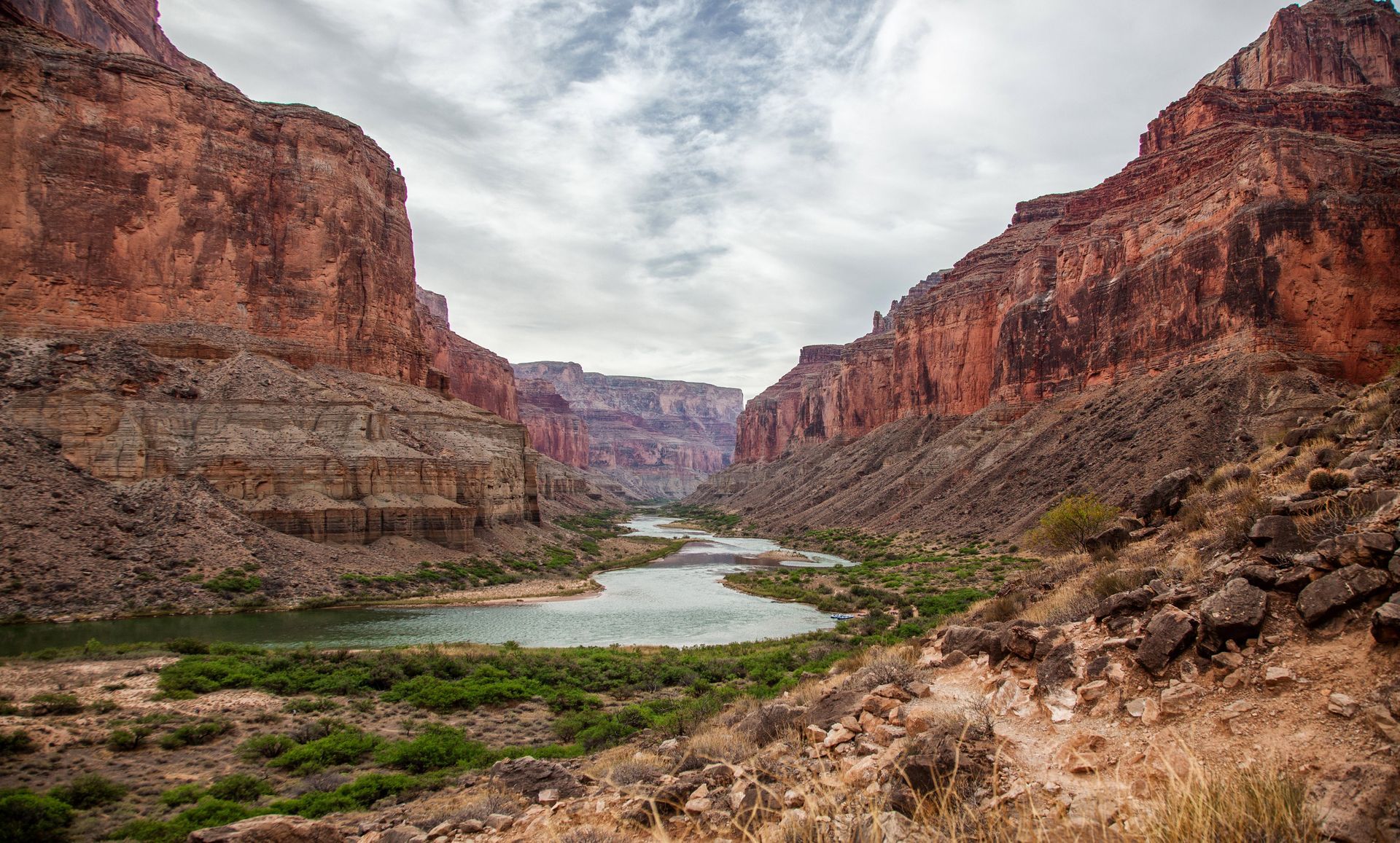 Canyon landscape with a river flowing between tall, reddish-brown rock walls. Cloudy sky overhead.