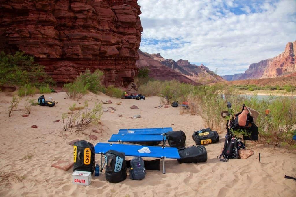 Camping gear on a sandy beach near a red rock wall and river under a cloudy sky.