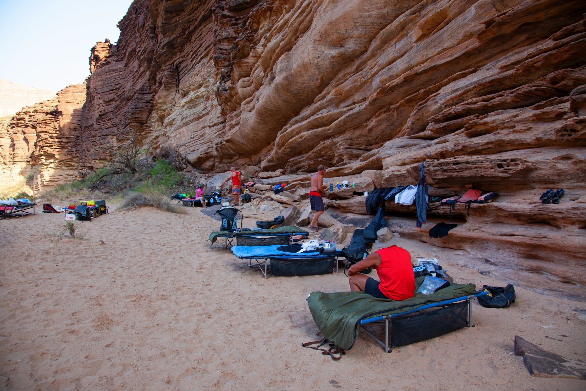 Campers relaxing on a sandy beach below a layered rock cliff.