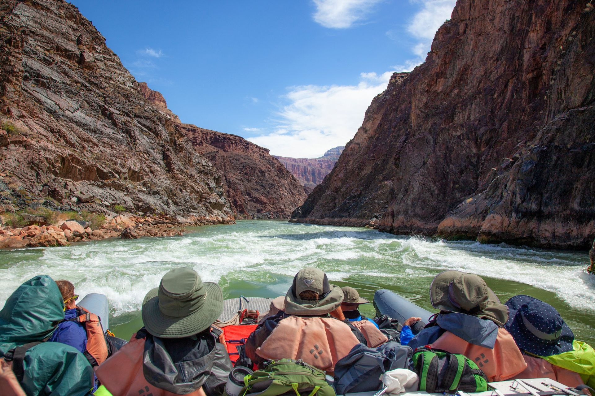 Rafting through a canyon; turbulent water, passengers wearing hats, cliffs on either side, blue sky above.