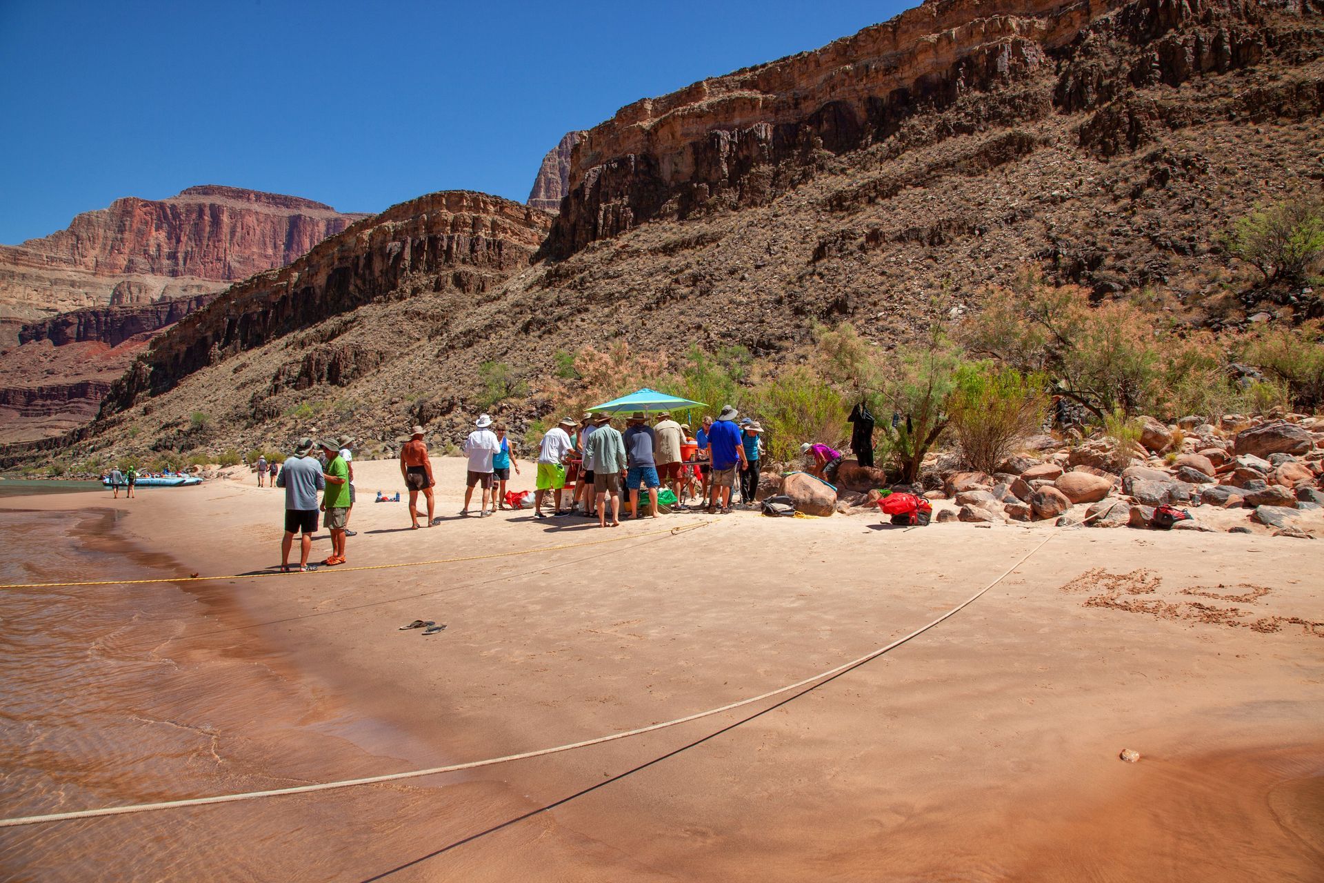 People gather on a sandy beach along a river, with red rock canyon cliffs in the background on a sunny day.