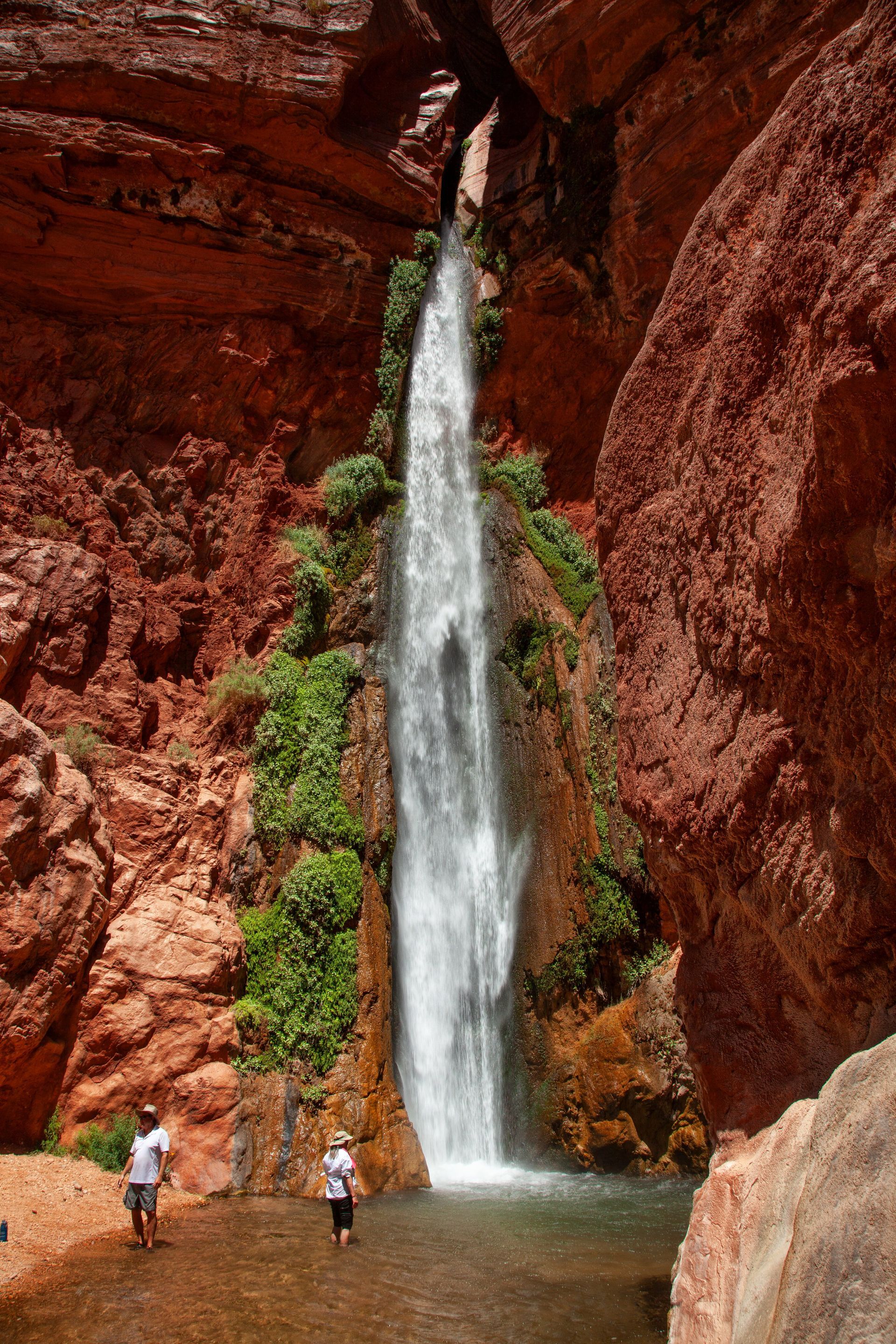 Waterfall cascading into a pool, flanked by red rock cliffs. Two people stand near the water.